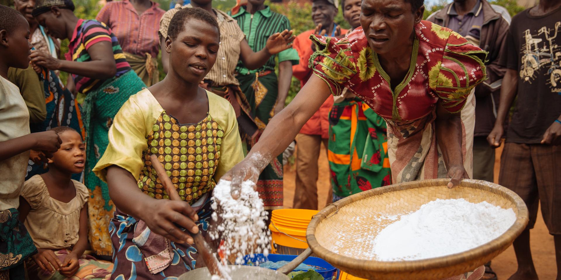 Woman hand sifting flour into a pot as another stirs with neighbors surrounding in a village outside of Lilongwe, Malawi