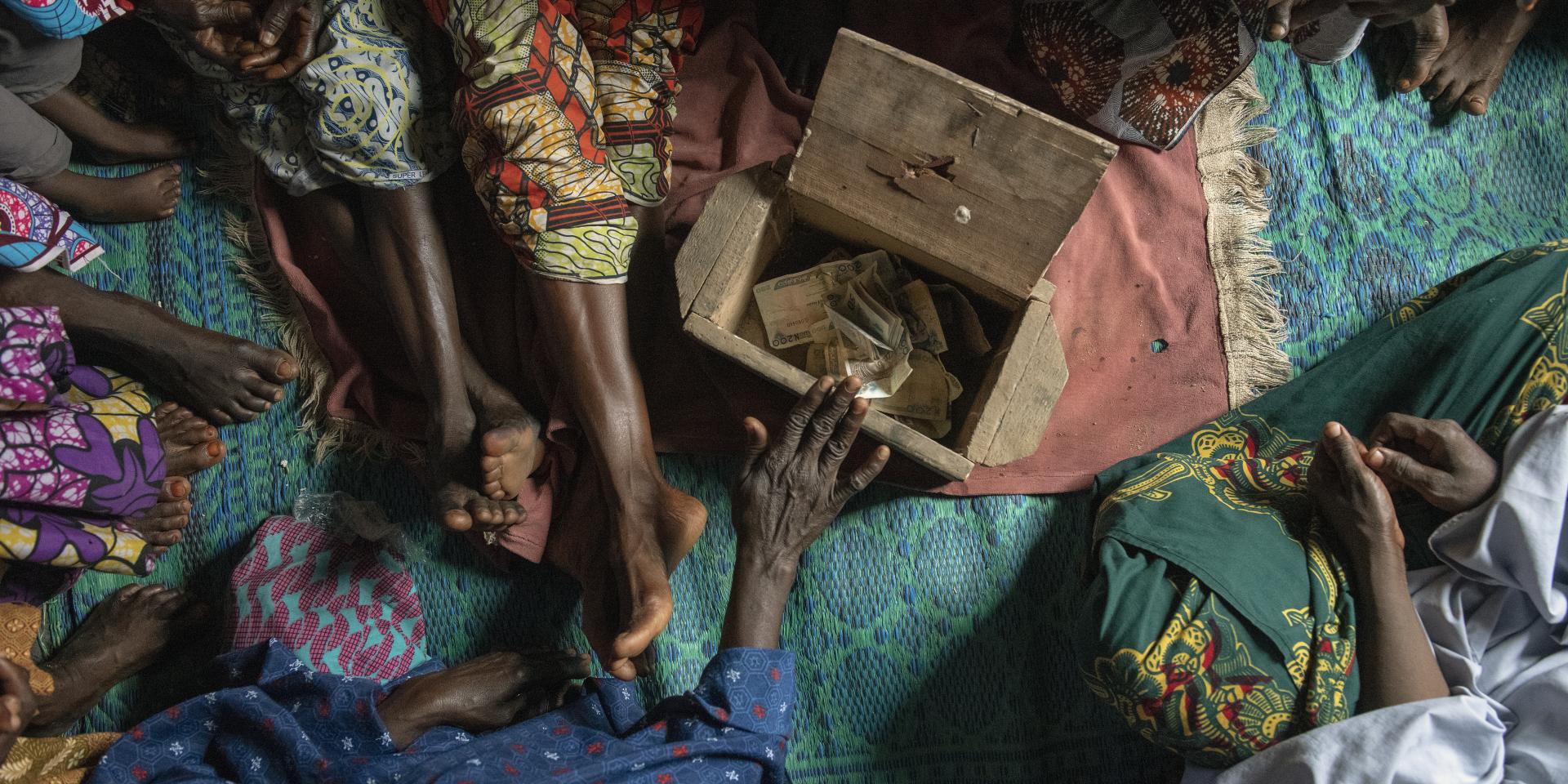 Women's hands place money into a shared savings box made of wood on a floor covered in blankets