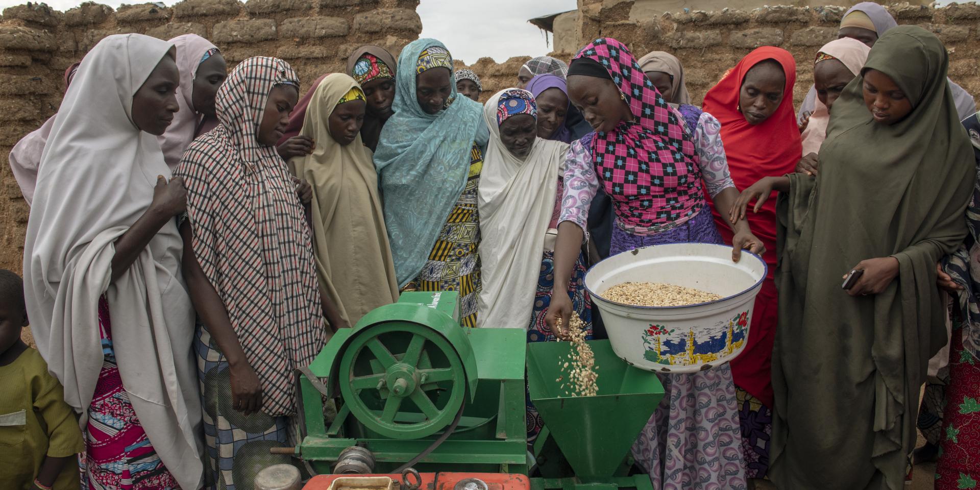 Women surround another woman with a bowl of groundnut standing next to a green machine as she places groundnut into the hopper