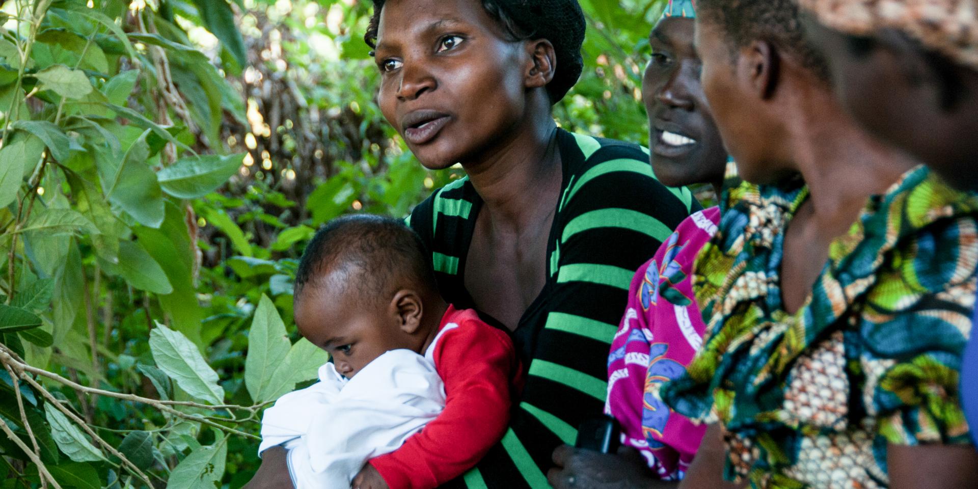 Women talking in shade near trees; one holds a baby