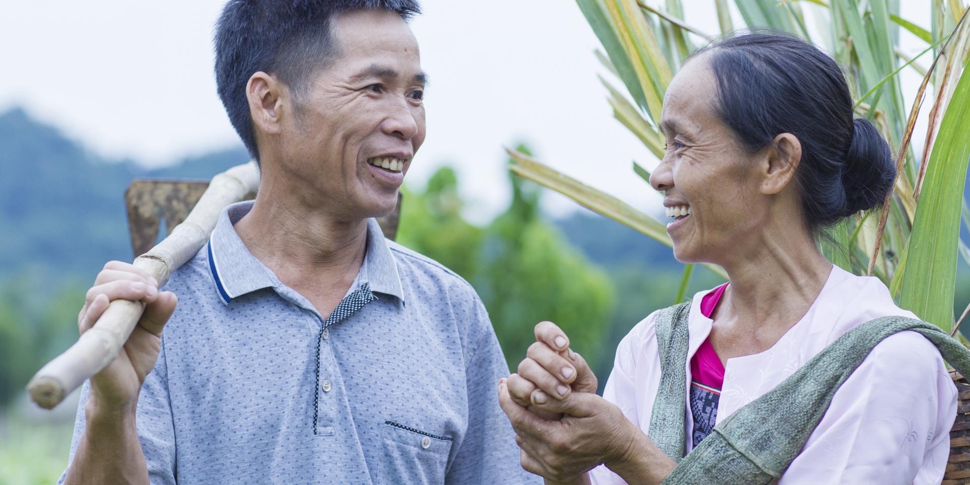 A man and woman look at each other in a field. One holds a tool. Muong ethnic people grow rice and keep pigs, buffalos, chickens in their house to generate more incomes