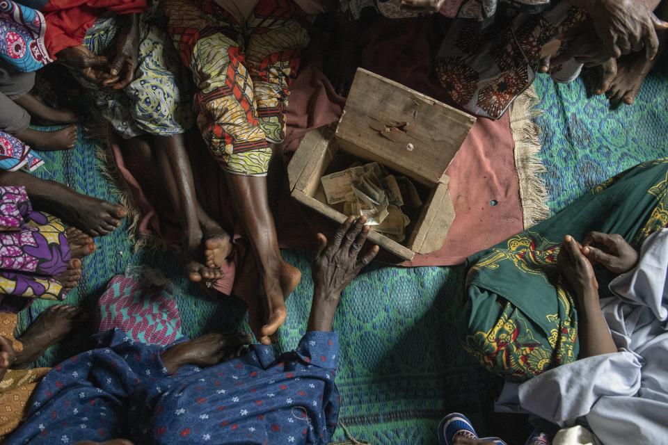 Women's hands place money into a shared savings box made of wood on a floor covered in blankets