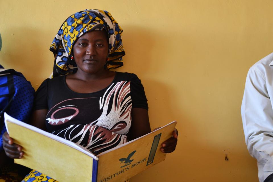 A woman looking at the camera sits in a building reading a book that says 'visitors book'