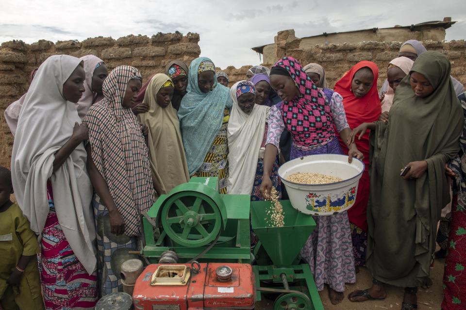 Women surround another woman with a bowl of groundnut standing next to a green machine as she places groundnut into the hopper