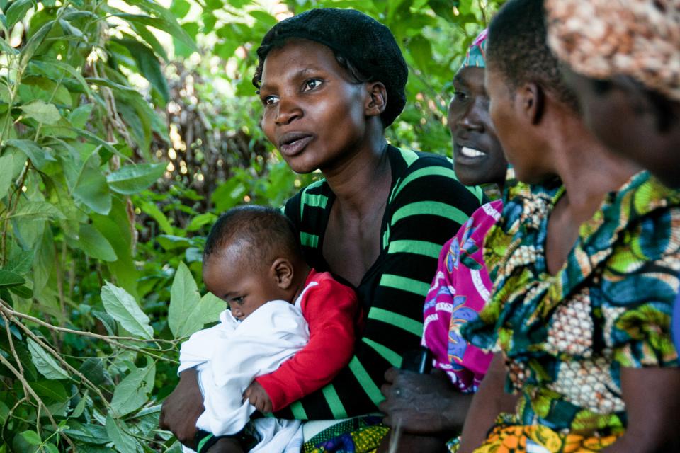 Women talking in shade near trees; one holds a baby