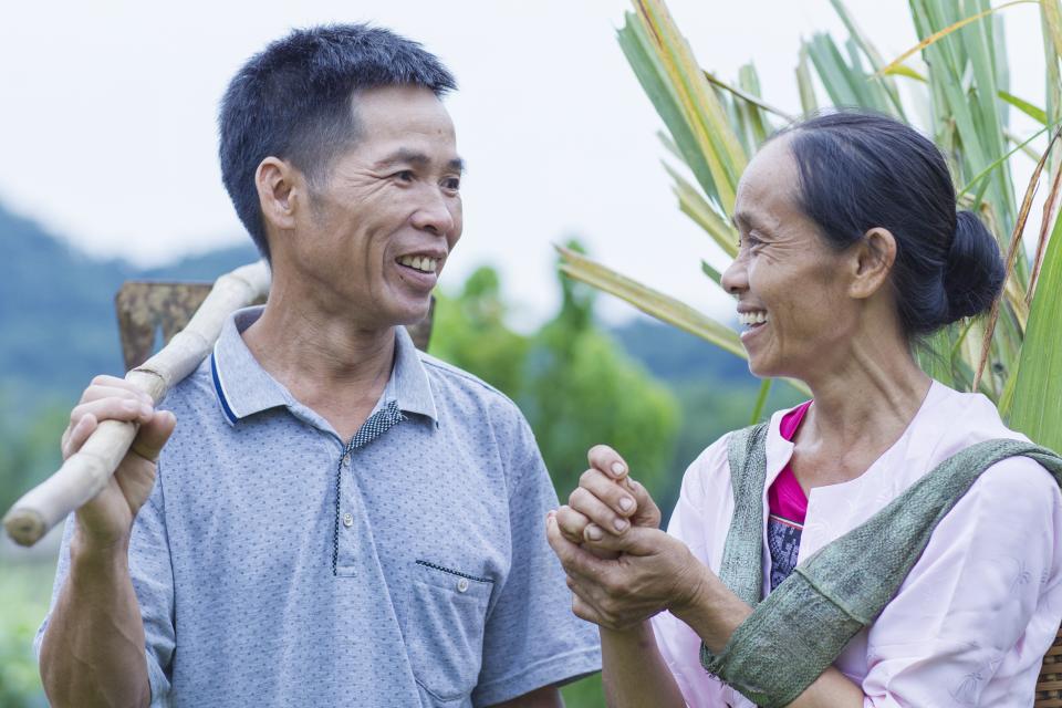 A man and woman look at each other in a field. One holds a tool. Muong ethnic people grow rice and keep pigs, buffalos, chickens in their house to generate more incomes