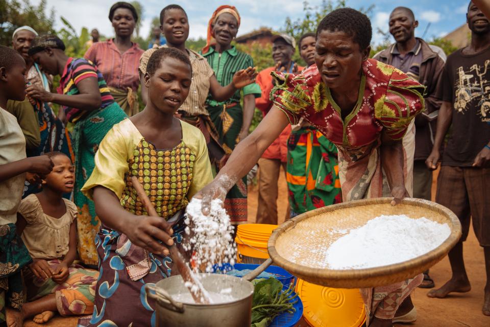 Woman hand sifting flour into a pot as another stirs with neighbors surrounding in a village outside of Lilongwe, Malawi