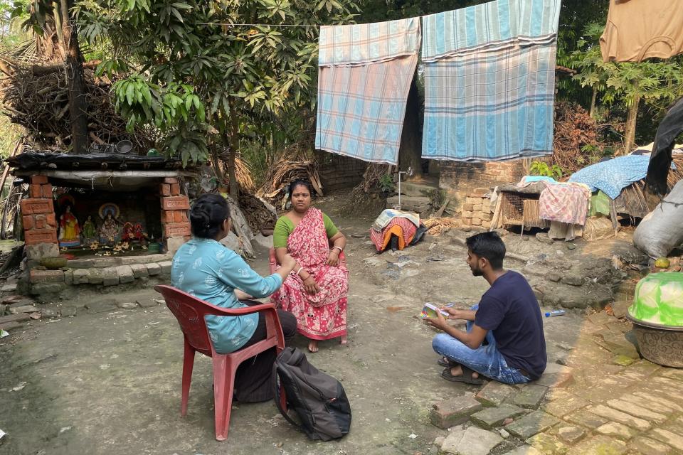 Two people sitting on chairs, one on the ground, outside with laundry drying in the background in West Bengal